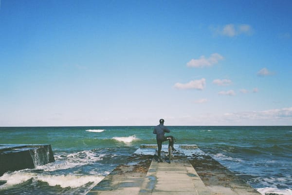 Lukasz Kus looking at the Baltic Sea, hands on camera, ready to take a photo,holding his bike with one one leg propped up on the seat